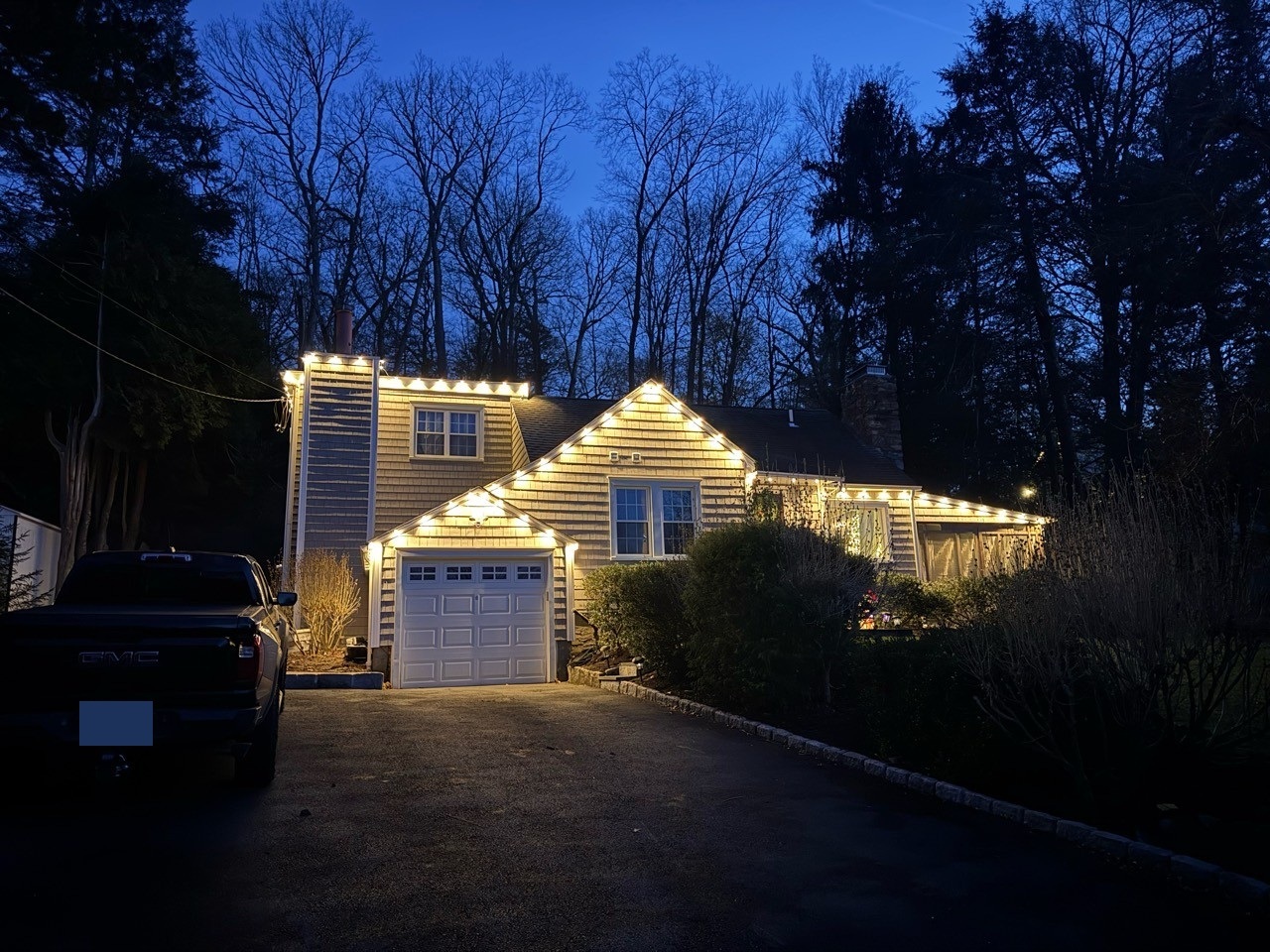 Warm white roofline lights across the garage, peaks and side of a shingled home at dusk